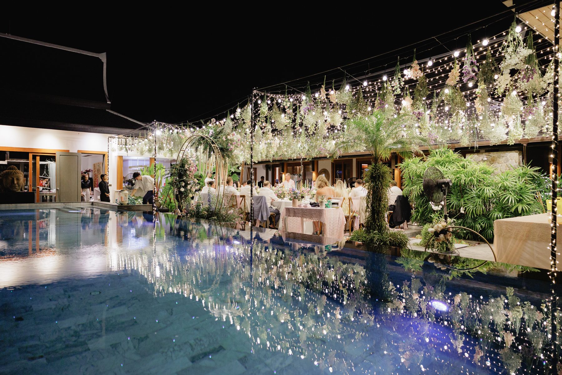 Long candlelit reception table beside the pool at Baan Phu Prana, dressed with low floral runners, by Wedding Flowers Phuket