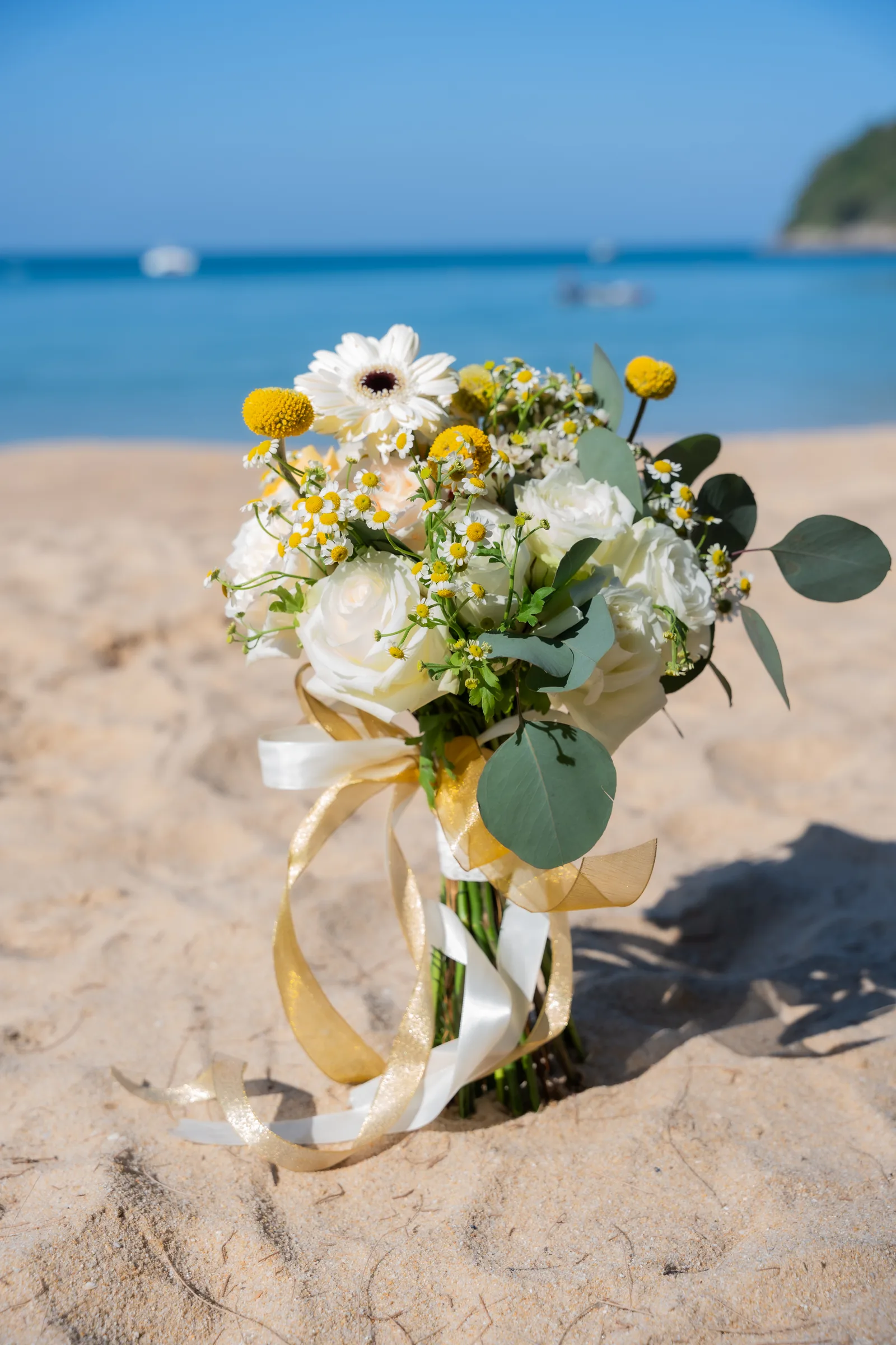 White and yellow bridal bouquet with chamomile, ranunculus and gold ribbon on Phuket sand